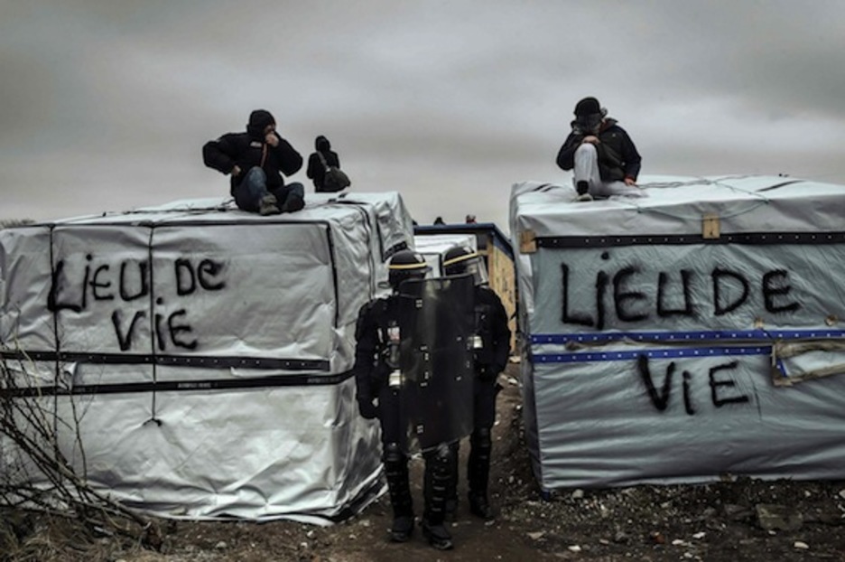 Policías pasan junto a migrantes y refugiados situados en los techos de las tiendas de Calais. (Philippe HUGUEN/AFP)