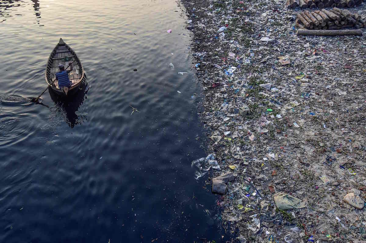 Gizon bat txalupa batean arraun egiten, plastikozko poltsak Dhakako Buriganga ibaiaren ur azalean flotatzen ari diren bitartean. (Munir UZ ZAMAN / AFP) 