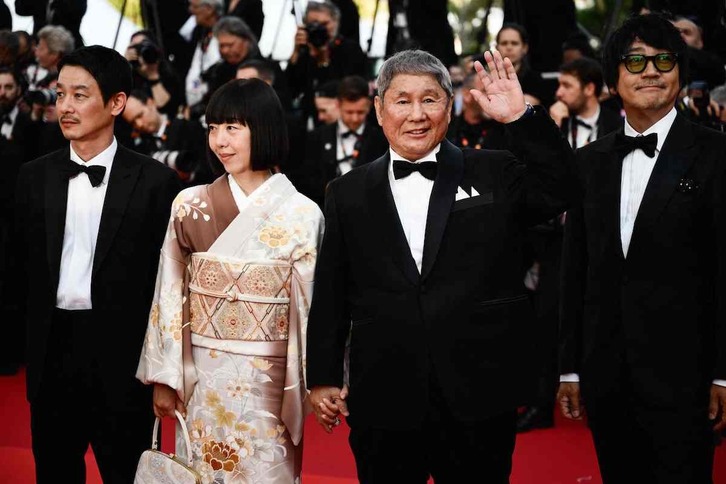 Takeshi Kitano saluda a los fotógrafos, acompañado por parte de su equipo.