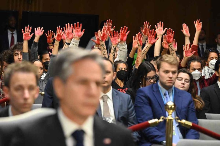 Los manifestantes levantan sus manos embadurnadas de pintura roja durante el discurso de Antony Blinken, en primer plano.